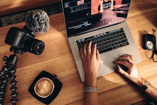 op view of female vlogger editing video on laptop. Young woman working on computer with coffee and cameras on table.