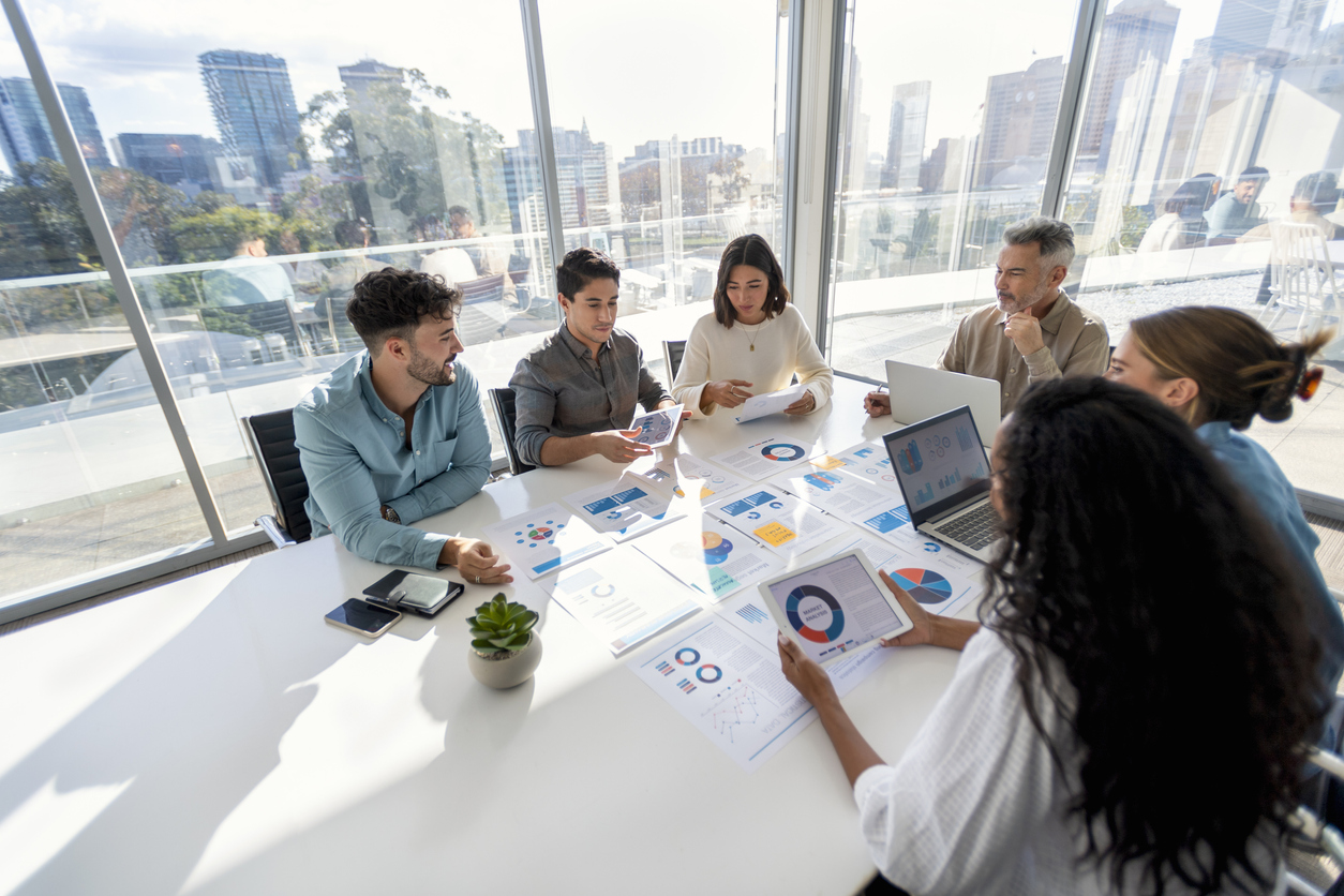 Multi racial group of people working with Paperwork on a board room table at a business presentation or seminar. The documents have financial or marketing figures, graphs and charts on them. Use data and surveys to connect with leaders on the job and promote your work with stats in a more powerful way.