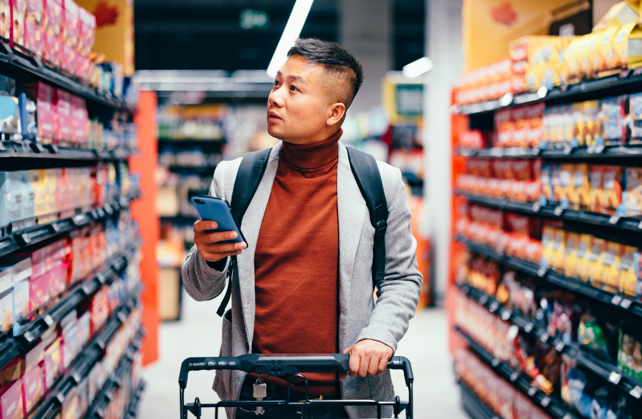 Male walking down the product aisle in the supermarket, looking at shelves and searching for groceries from the list on his mobile phone he is holding in his hand. As the recession fears are uncertain people are scaling back on spending money.