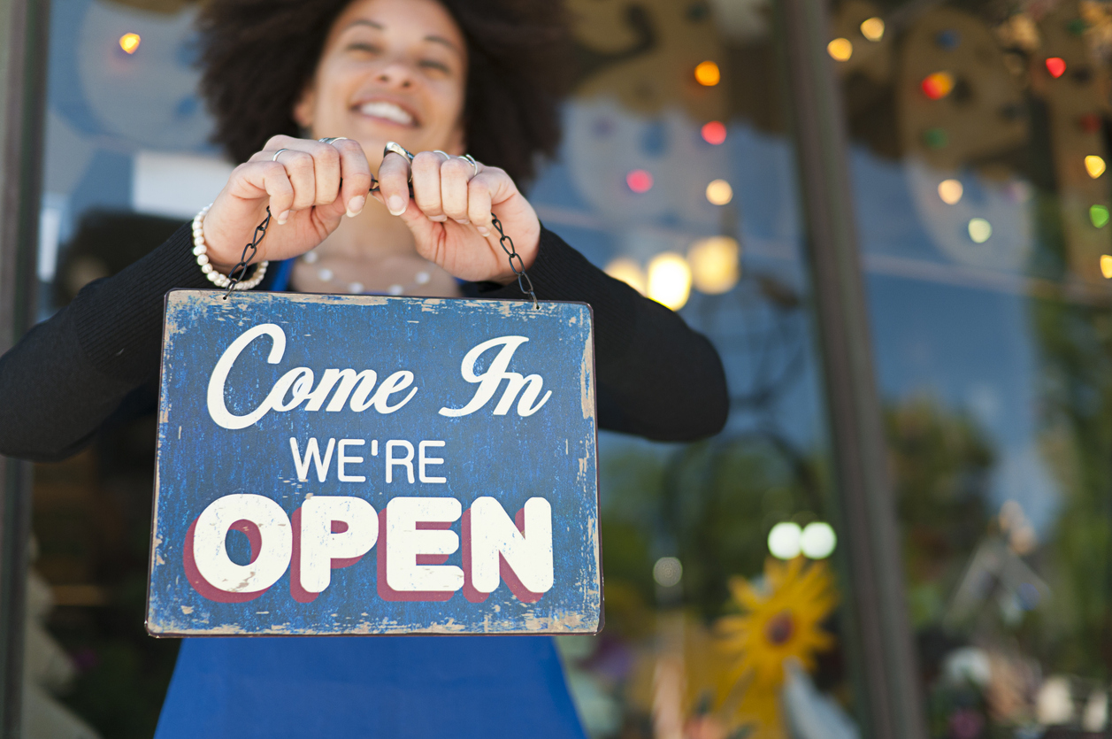 Woman holding vintage open sign up outside of a boutique store front. Brands can learn new campaign ideas to support Black Business Month.