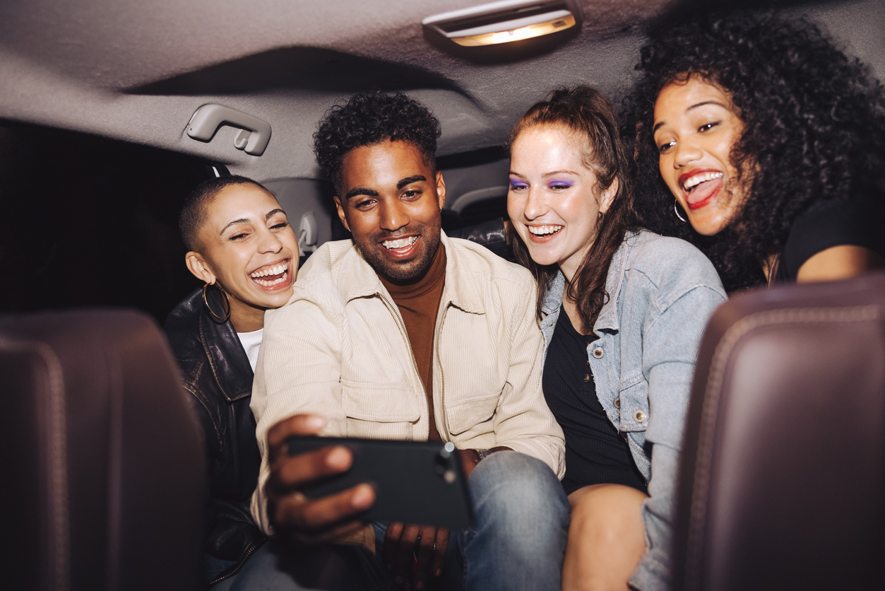 Happy friends taking a selfie together inside a car at night. Group of young friends smiling cheerfully while posing for a group photo. Carefree friends taking a ride home after a party. Uber, Madd and Anheuser-Busch are joining forces to connect with college students now to encourage safe riding in a new campaign.