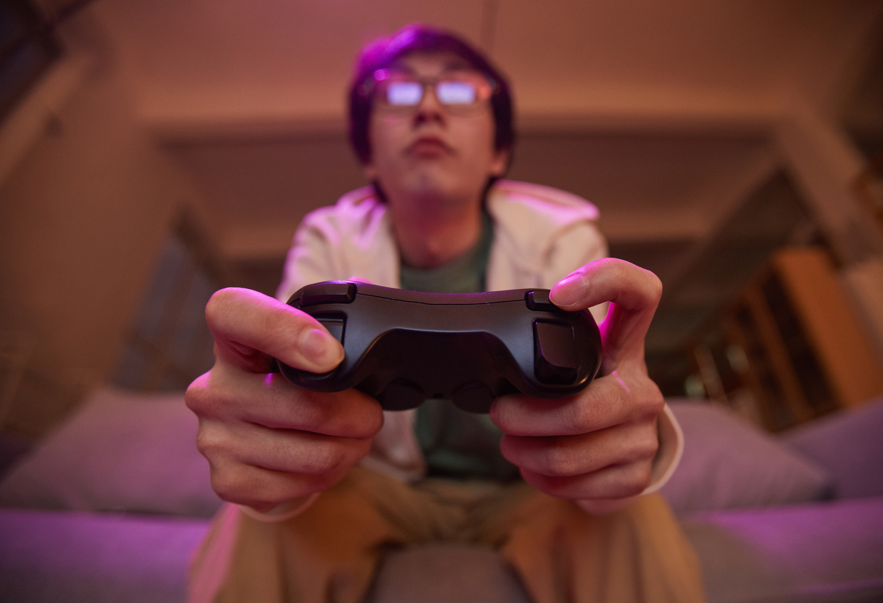 Low angle view at Asian young man holding gamepad and playing video games at home lit by purple light, copy space. Atari is building its brand with more throwback nostalgic products like Atari 50.